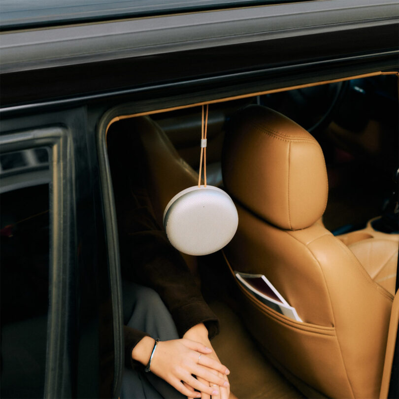 A Beosound A1 portable speaker hangs from the top of a car window next to a person seated inside, with tan leather seats and a tablet in the seat pocket.