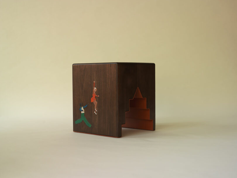 A wooden side table featuring a cut-out stepped design and an inlaid illustration of two people reaching upward on one side.