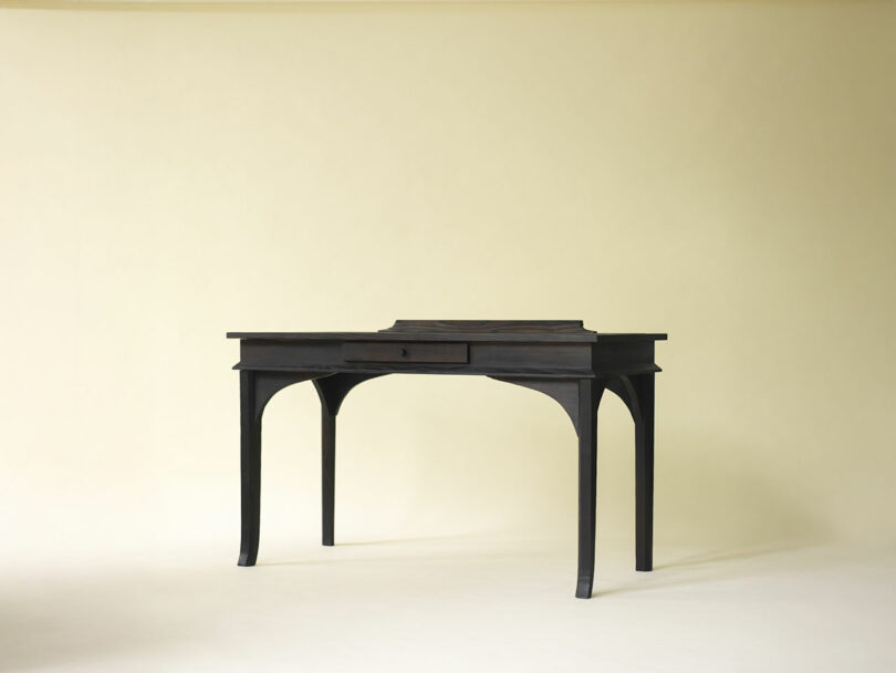 A dark wooden desk with curved legs, a single central drawer, and a raised back section, set against a plain light background.