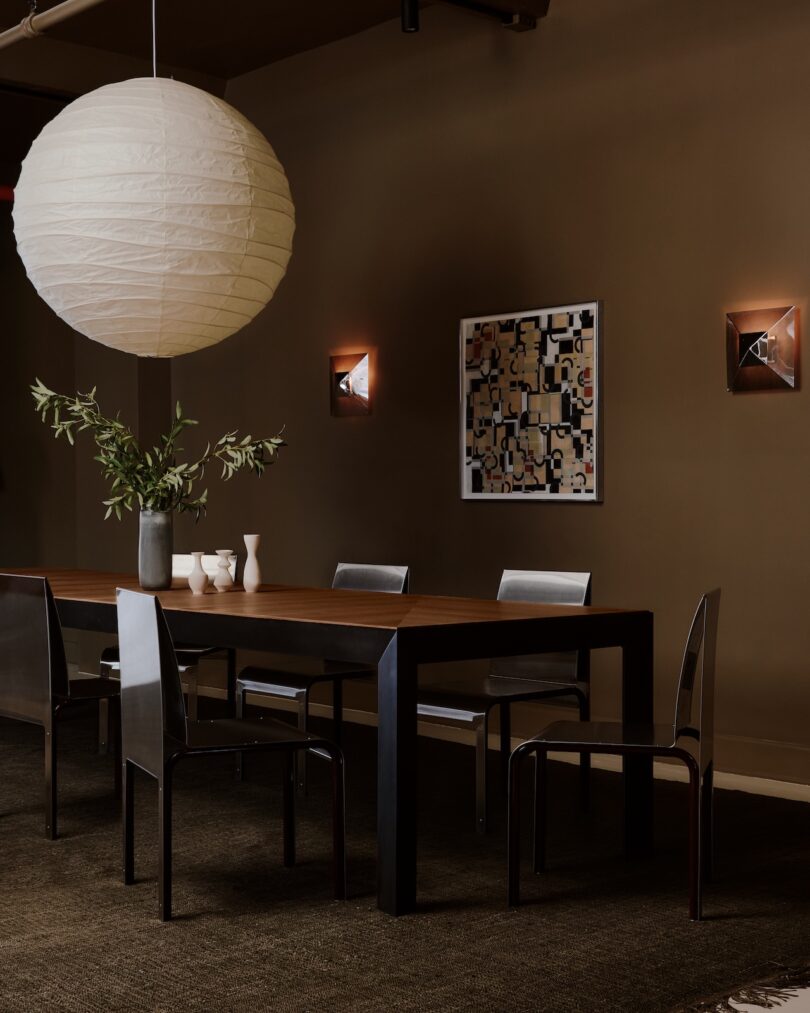 A modern dining room with a long wooden table, six black chairs, a large round paper lantern, wall art, and a vase with greenery.