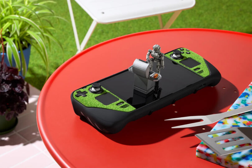 A green handheld gaming console sits on a red table outdoors with a small robot figurine and gray stand placed on its screen.