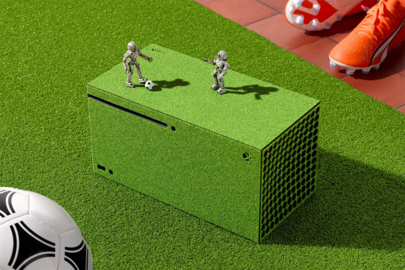 A green Xbox console resembling grass with two small robot figures playing soccer on top, next to soccer cleats and a soccer ball on artificial turf.