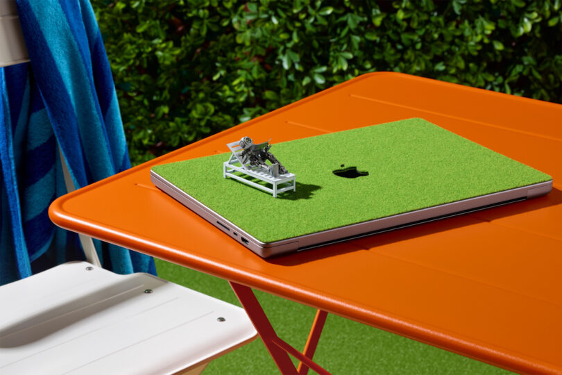 A closed laptop with a green grass-like cover and a miniature lounge chair figurine sits on an orange outdoor table.