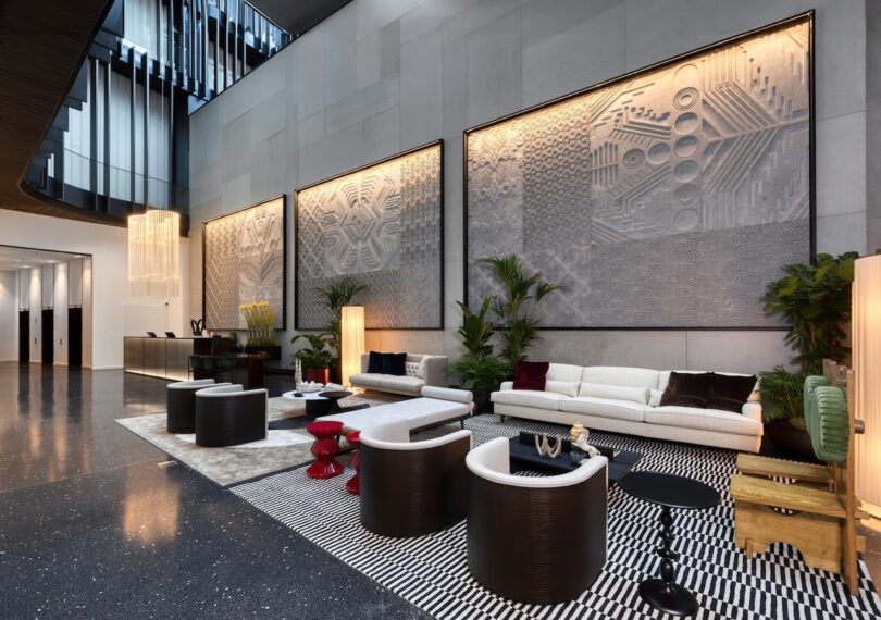 Modern lobby with contemporary furniture, large textured wall art, indoor plants, and a high ceiling with abundant natural light.