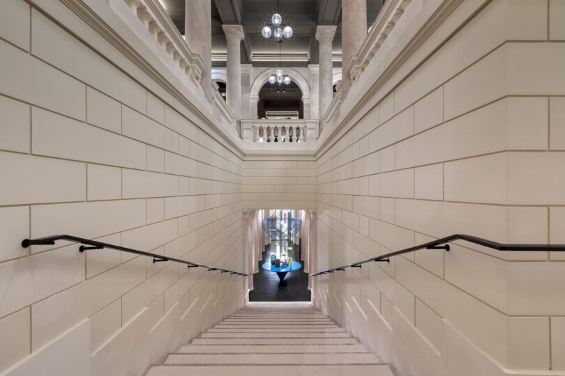 Wide staircase with cream-colored walls leads down to a modern area with blue chairs, beneath a grand architectural space with columns and hanging lights.