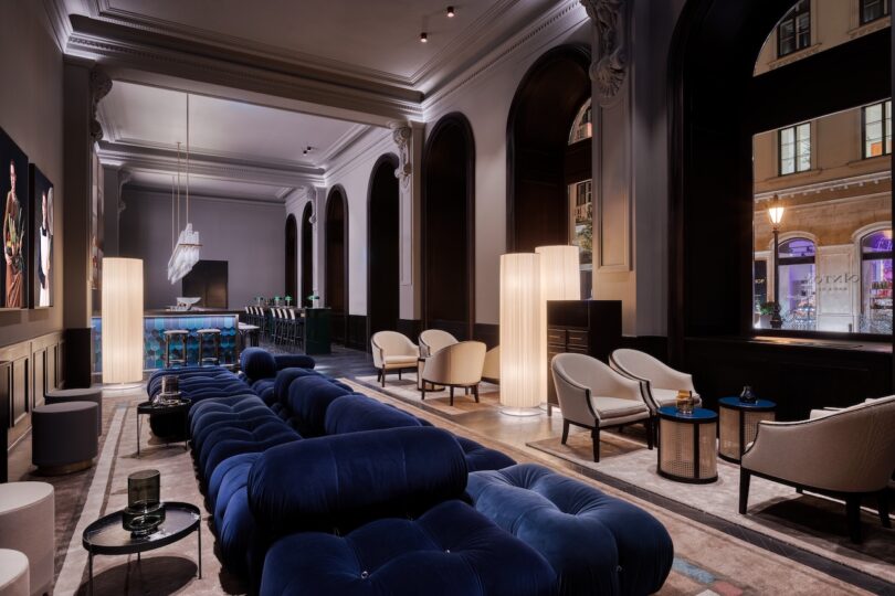 Modern hotel lounge with deep blue sofas, cream armchairs, large arched windows, tall floor lamps, and a bar area in the background.