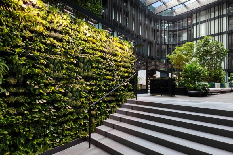 Modern indoor atrium with a green living wall, large potted plants, a stairway with metal railing, and natural light streaming through a glass ceiling.