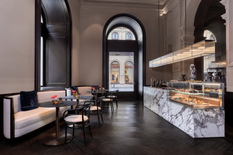 Modern cafe interior with marble counter, glass display case of pastries, black tables and chairs, and large arched window showing shops outside.