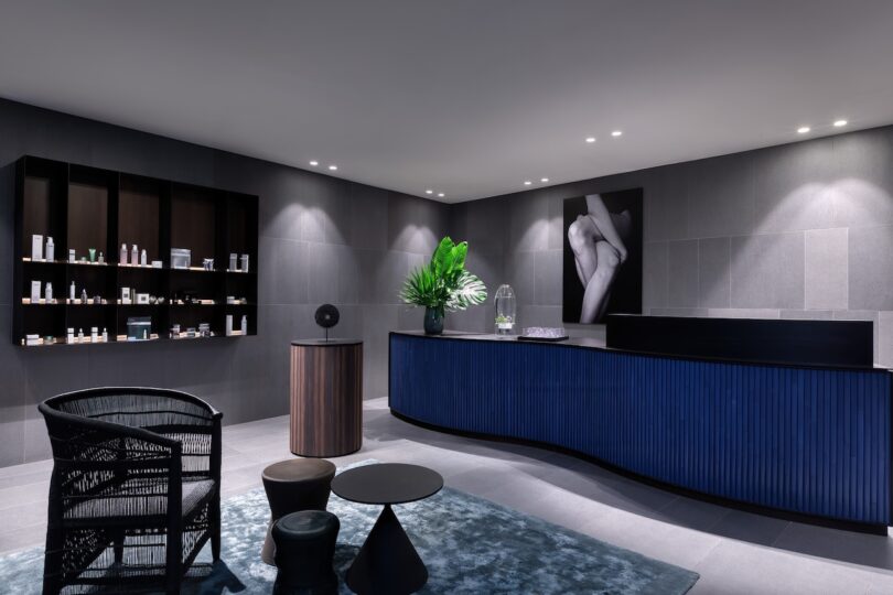 Modern spa reception area with a blue front desk, display shelves of products, black chair, small tables, a green plant arrangement, and a black-and-white artwork on gray tiled walls.