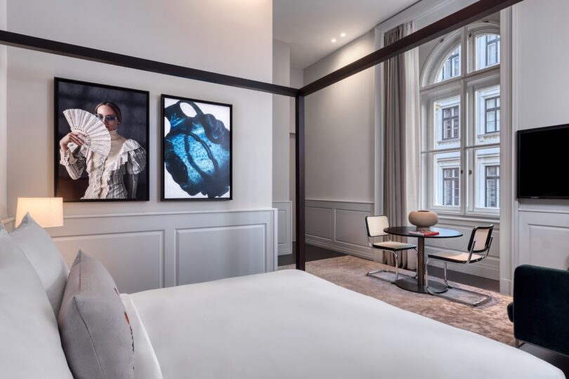 A modern hotel room with a four-poster bed, two framed artworks on the wall, a round table with two chairs, and large windows with city views.