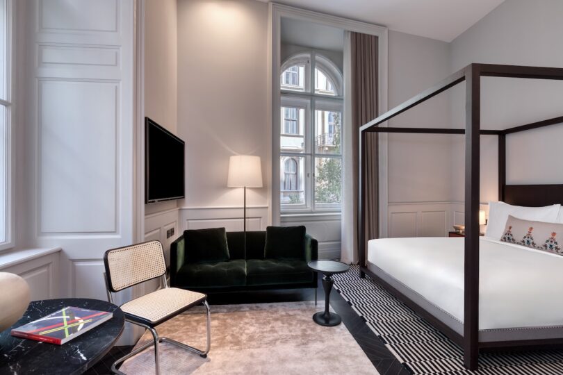 Modern hotel room with a four-poster bed, dark green sofa, chair, round table, large window, and minimalist decor in neutral tones.