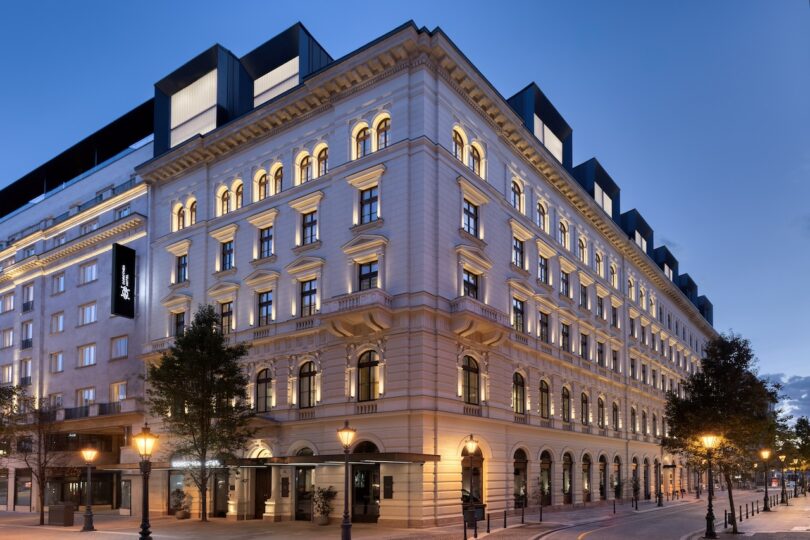 A large, elegant, multi-story hotel building with illuminated windows stands on a corner street at dusk, featuring classic architectural details and street lamps.