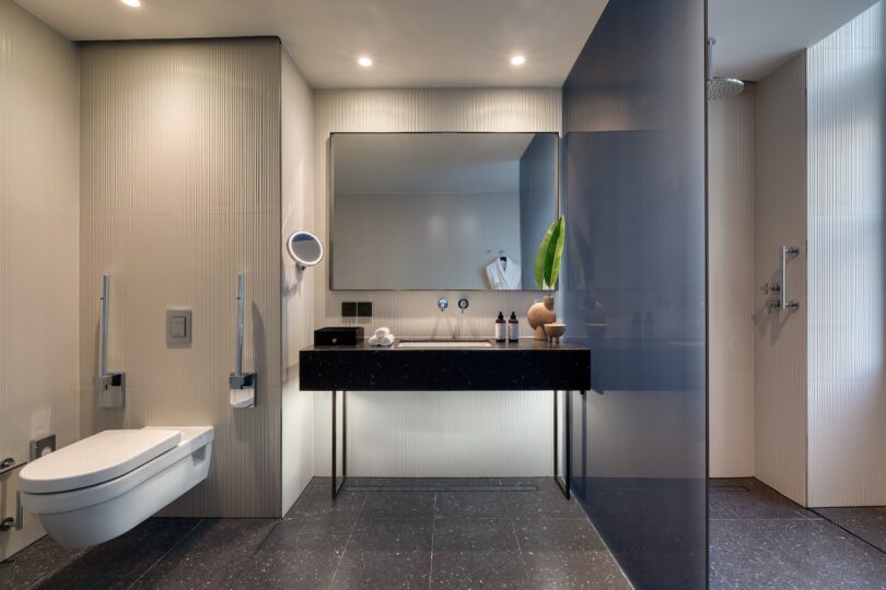 Modern bathroom with a wall-mounted toilet, large mirror above a black floating vanity, various toiletries, and a walk-in shower with glass partition.