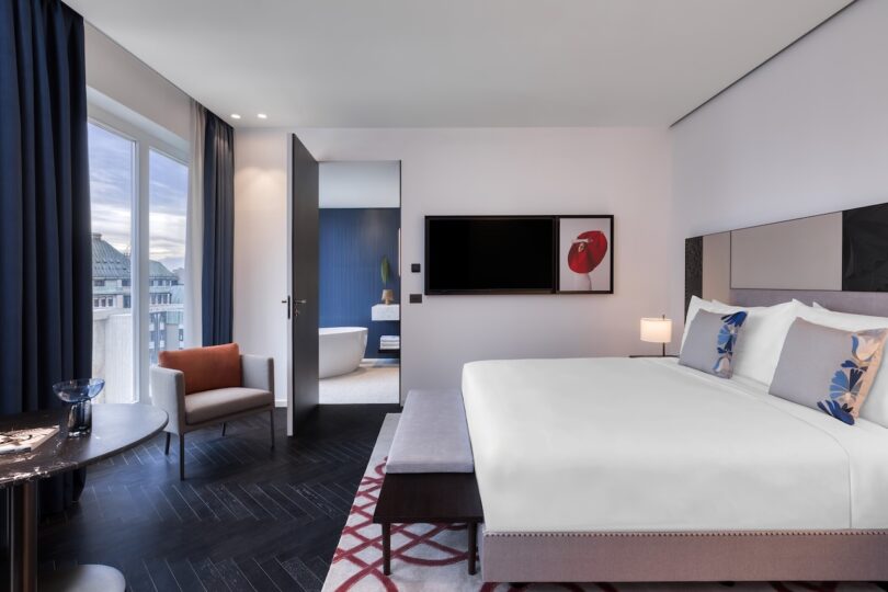 Modern hotel room with a large bed, TV, armchair, table, and window; open door reveals a bathroom with a freestanding tub.