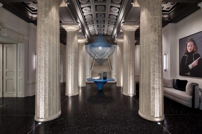 A modern interior features tall, fluted columns, a blue geometric table centerpiece, a floating chandelier, black speckled floors, and a portrait of a woman on the right wall.