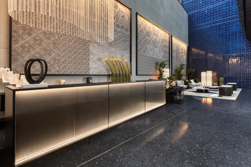 Modern lobby with a sleek reception desk, large textured wall art, decorative vases, and a lounge area with contemporary seating and lighting.