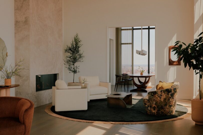 Modern living room with neutral-toned furniture, a round black rug, indoor plants, wall-mounted fireplace, and adjoining dining area with large windows.