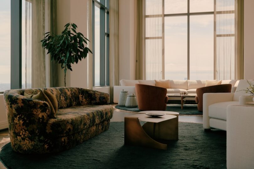 A modern living room with large windows, a green floral sofa, two brown armchairs, a white sofa, a black rug, and a potted plant.