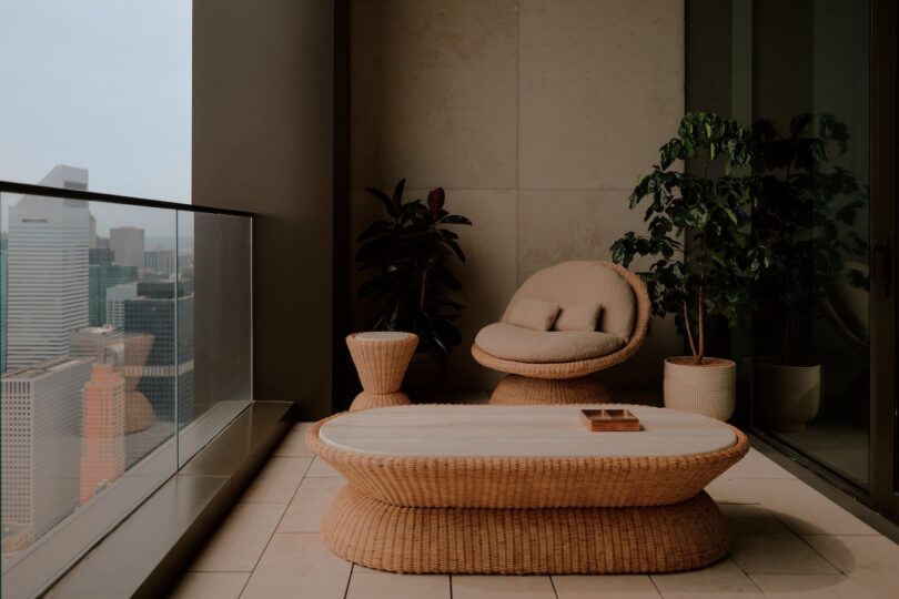 A modern balcony with wicker furniture, including a table, chair, and side table, potted plants, and a cityscape view through glass railing.