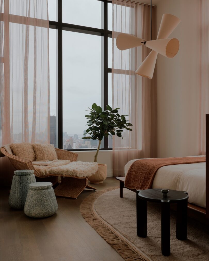 A modern bedroom with large windows, sheer pink curtains, a bed with brown bedding, a lounge chair with a throw, a potted plant, and various small tables.