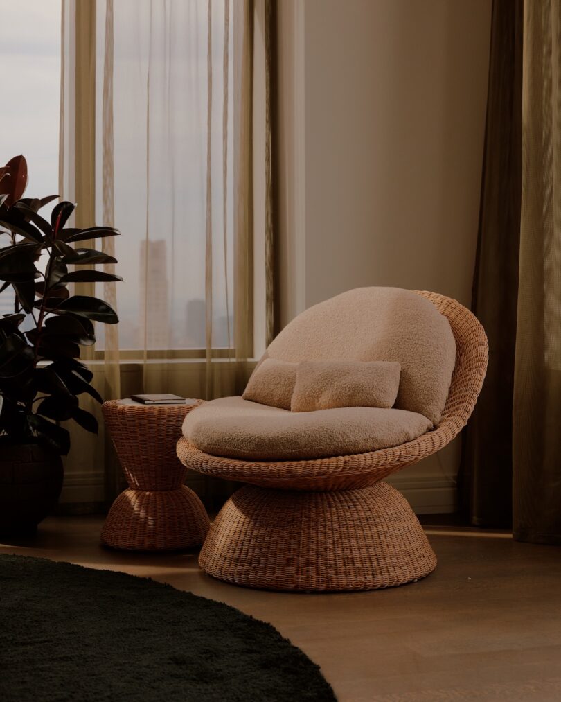 A round wicker chair with beige cushions and a matching side table sit by a window with sheer curtains in a softly lit room.