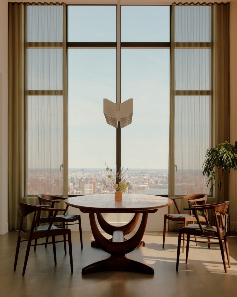 A modern dining area with a round wooden table, six chairs, a potted plant, and large windows overlooking a cityscape.