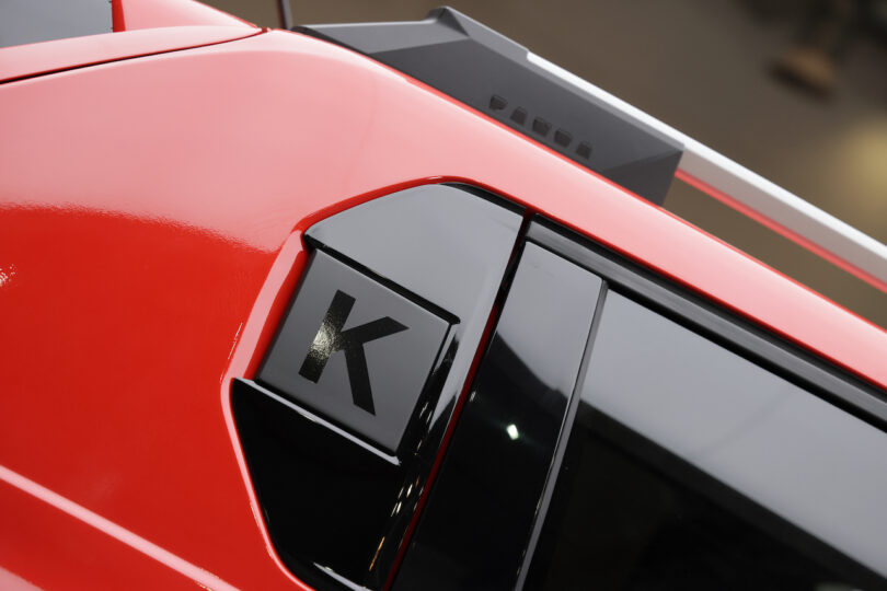 Close-up of a red sports car’s rear side window with a black "K" decal and a prominent angular rear spoiler