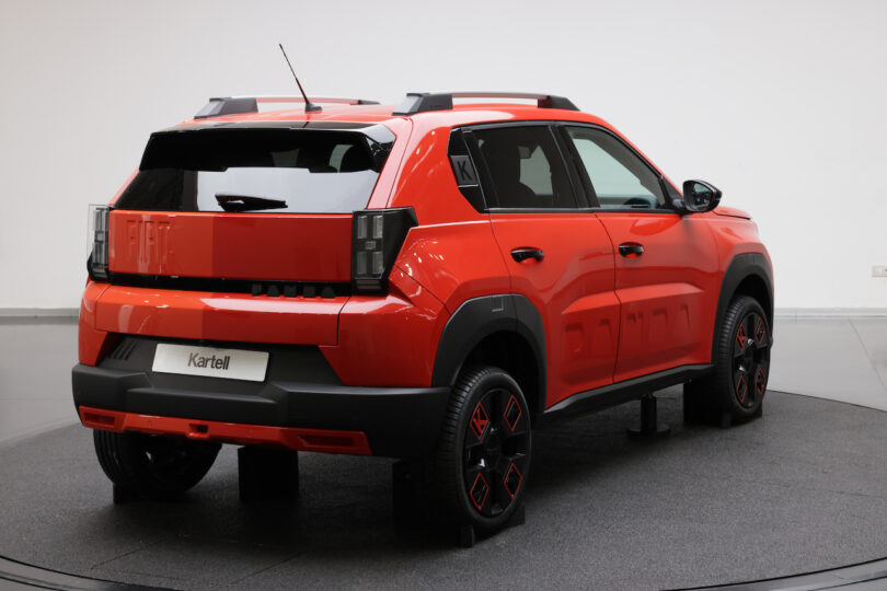 A red Fiat SUV is displayed indoors on a circular platform, viewed from the rear passenger side. The car features black accents, roof rails, and bold wheel designs
