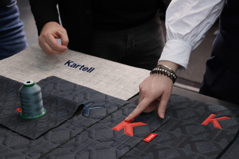 Close-up of hands arranging textile samples, thread spool, and red Kartell logo pieces on a table with "Kartell" text visible on fabric in the background