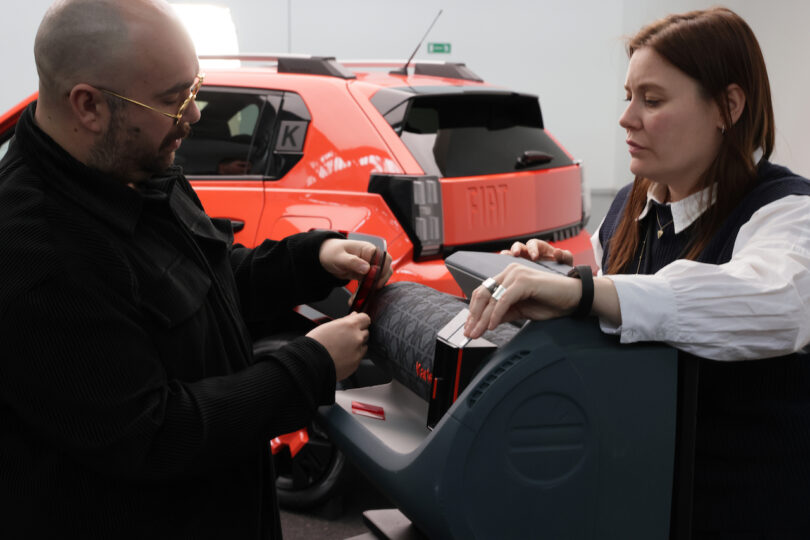 Two people examining or adjusting material on a machine, with an orange Fiat car visible in the background