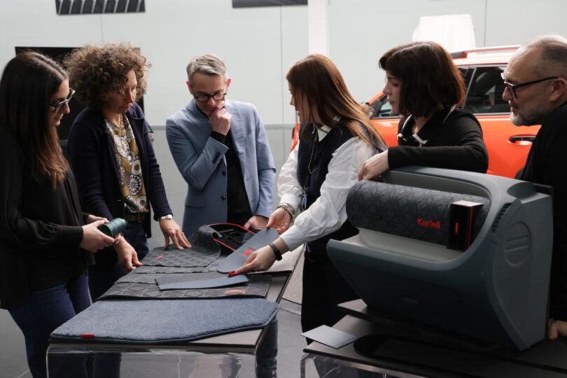 Six people stand around a table examining fabric samples and automotive materials, with a red car and a Kartell-branded machine in the background
