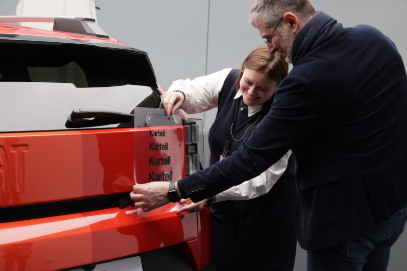 Two people attach a clear panel labeled "Kartell" to the rear side of a red vehicle