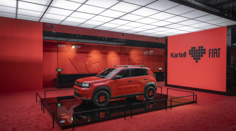 A red Fiat SUV is displayed on a raised platform in a showroom with red walls and flooring, featuring "Kartell ♥ FIAT" signage in the background