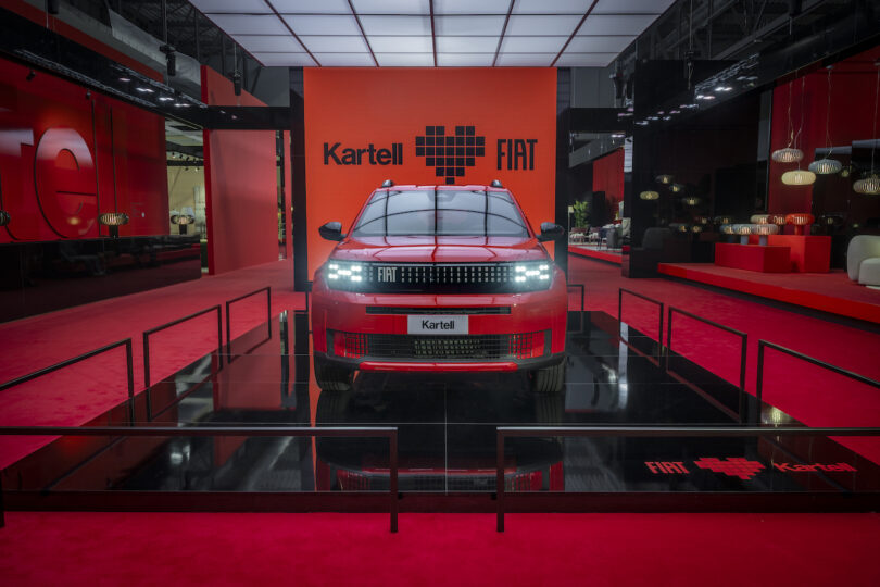 A red Fiat car is displayed on a black platform at an indoor event, with Kartell and Fiat logos visible on a red backdrop behind the vehicle