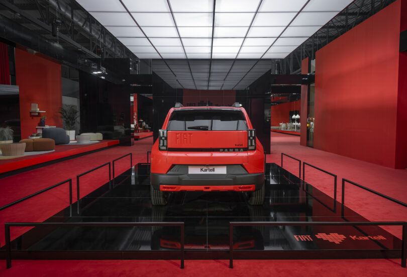 A red Fiat concept car is displayed on a black platform in a modern showroom with red floors and walls, surrounded by minimal black railings
