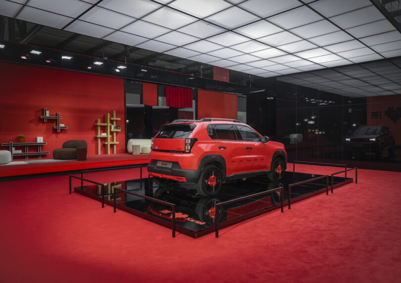A red SUV is displayed on a platform in a modern, red-themed showroom with contemporary furniture and reflective walls