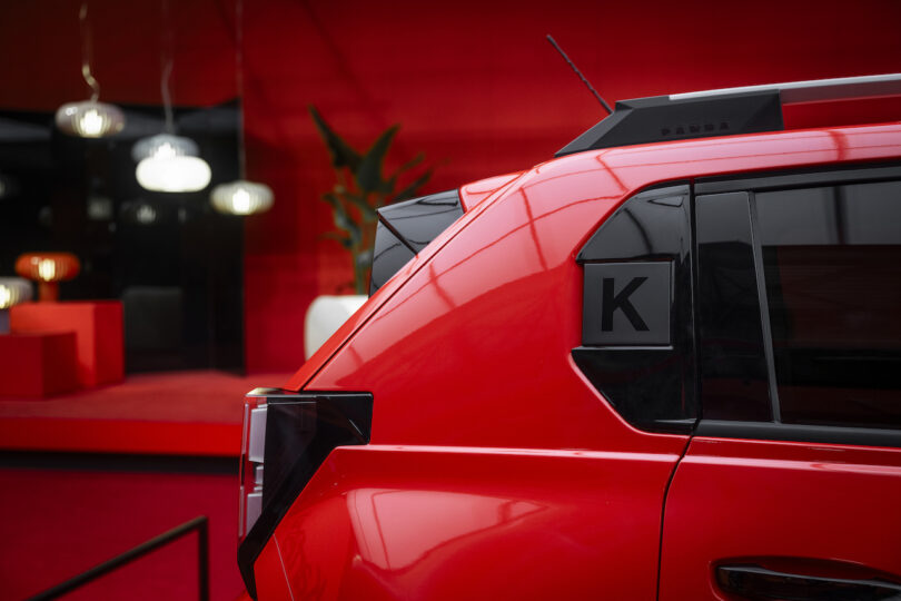 A close-up of the rear side of a red SUV with a black "K" decal on the window, set against a red background with modern furniture and pendant lights