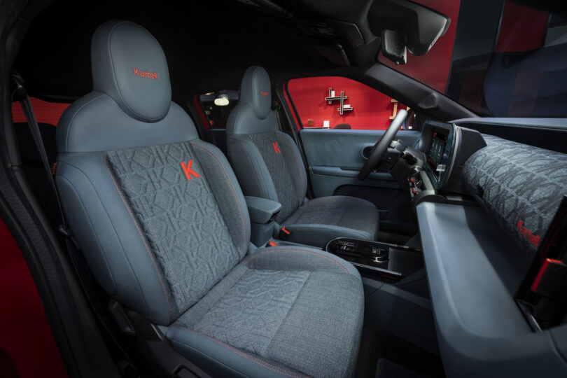 Interior view of a modern car featuring two front seats with gray upholstery, red accents, and embroidered letter "K" on the seats and headrests