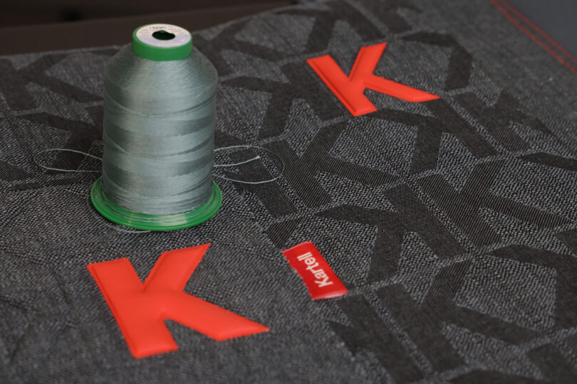 A spool of gray thread sits on dark fabric with red "K" logos and a small red Kanati label