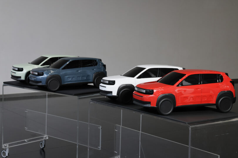 Four small SUV car models in matte green, blue, white, and red are displayed on transparent stands against a plain background