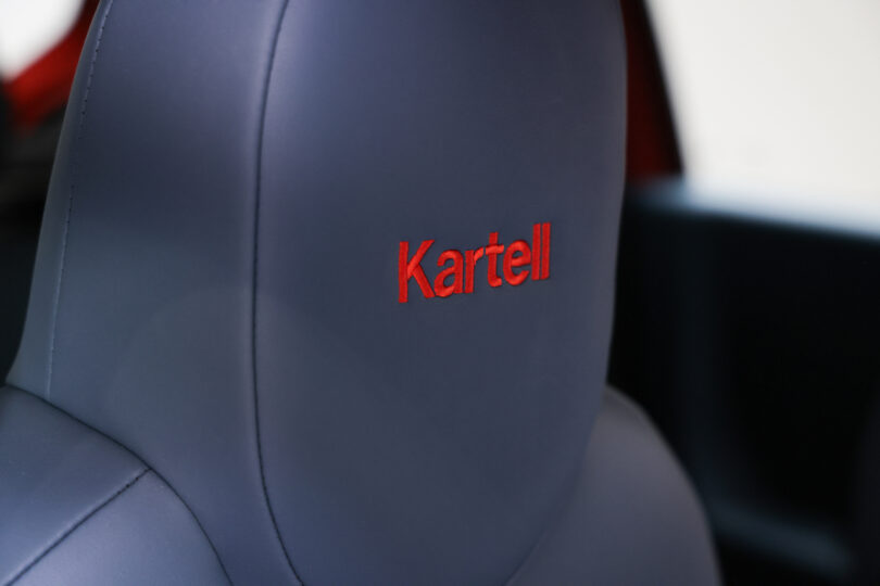 Close-up of a car seat headrest upholstered in dark material with the word "Kartell" embroidered in red on the back.