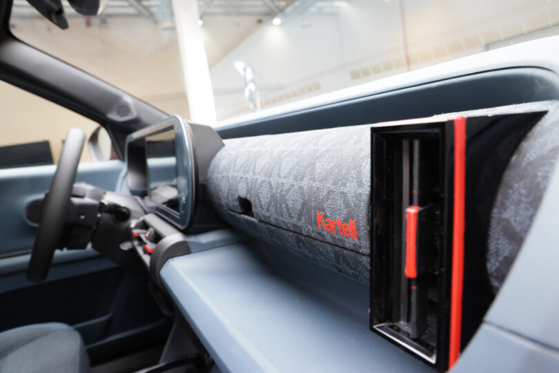 Close-up of a car dashboard featuring a fabric-covered panel with the word "Kartell," a screen near the steering wheel, and a rectangular air vent with a red accent