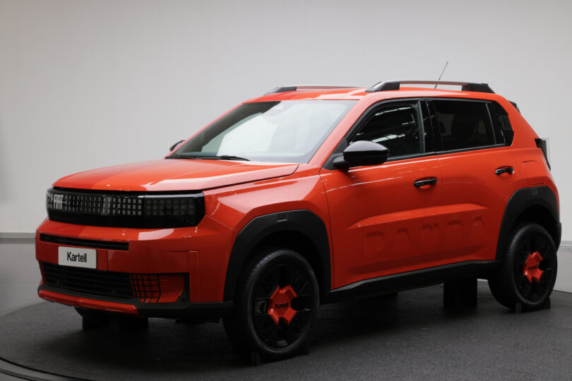 A bright orange compact SUV with black accents and red hubcaps, displayed indoors on a circular platform with a plain light background