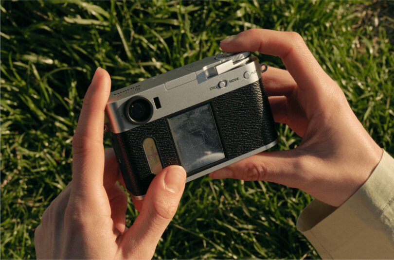 A person holds a compact, silver and black film camera with both hands, standing on grass in natural daylight.