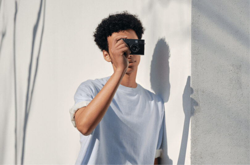 A person wearing a white t-shirt stands against a white wall, holding a camera up to their face and taking a photo.