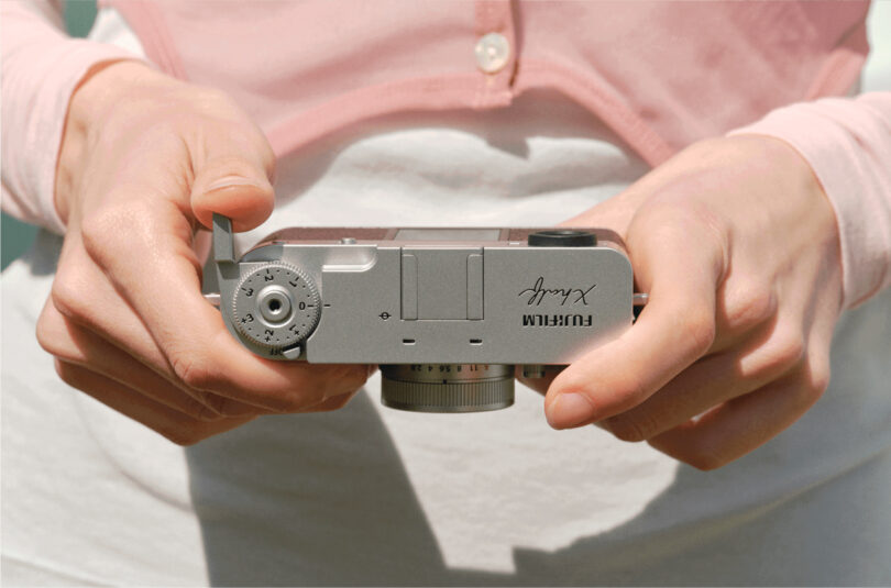 Close-up of a person holding a Fujifilm X100V camera, showing their hands and the top view of the camera with control dials visible.