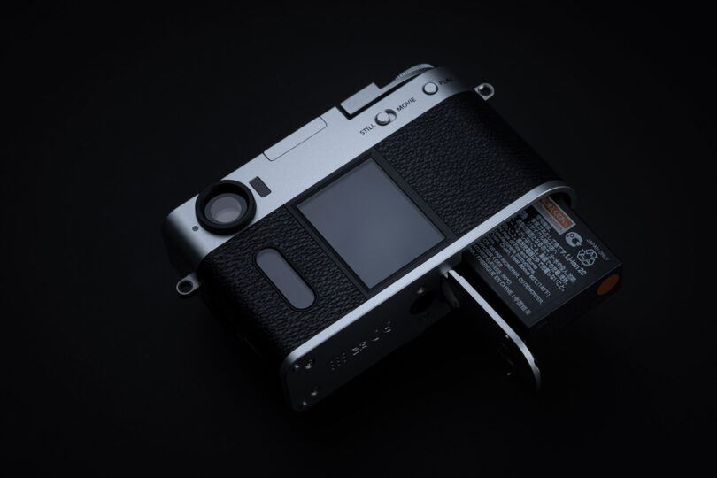 A digital camera with its battery compartment open and the battery partially ejected, displayed on a dark background.