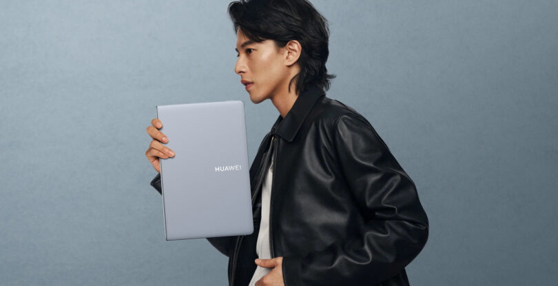 Person in a black leather jacket holding a silver Huawei laptop upright against a plain gray background.