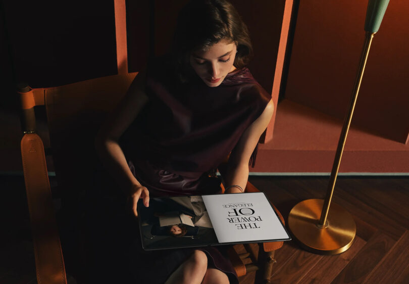 A person sits in a chair reading a magazine titled "The Power of Elegance" in a dimly lit room with a gold floor lamp and wooden floor.