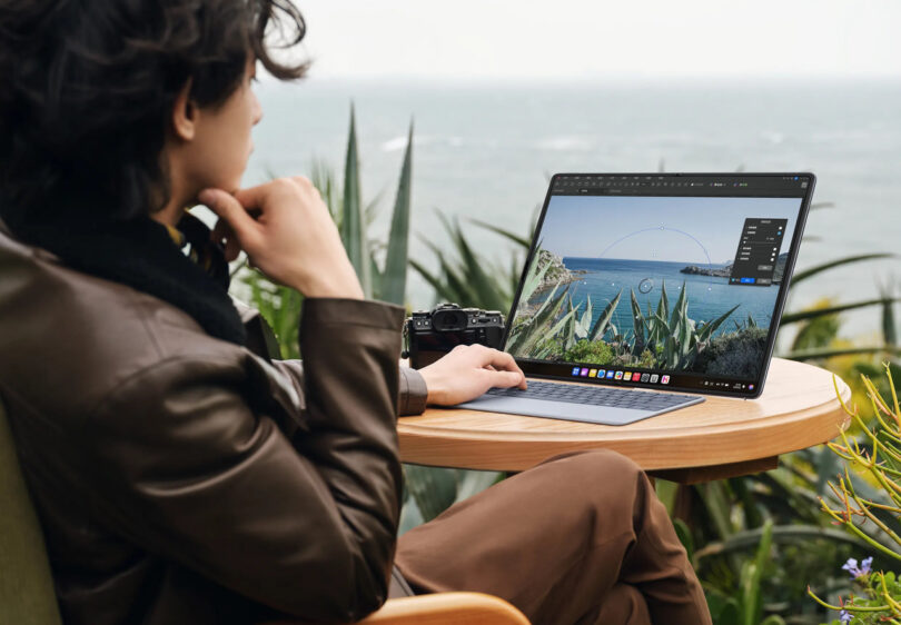 A person sits at an outdoor table by the sea, editing a photo of the ocean on a laptop; a camera rests on the table beside them.